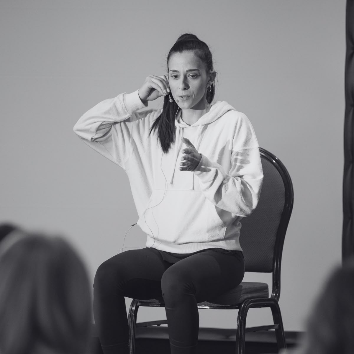 Ioanna laoumtzi καθισμένη σε καρέκλα μιλά με μικρόφωνο και κάνει χειρονομίες μπροστά σε κοινό. / Seated woman speaks into a microphone and gestures to an audience